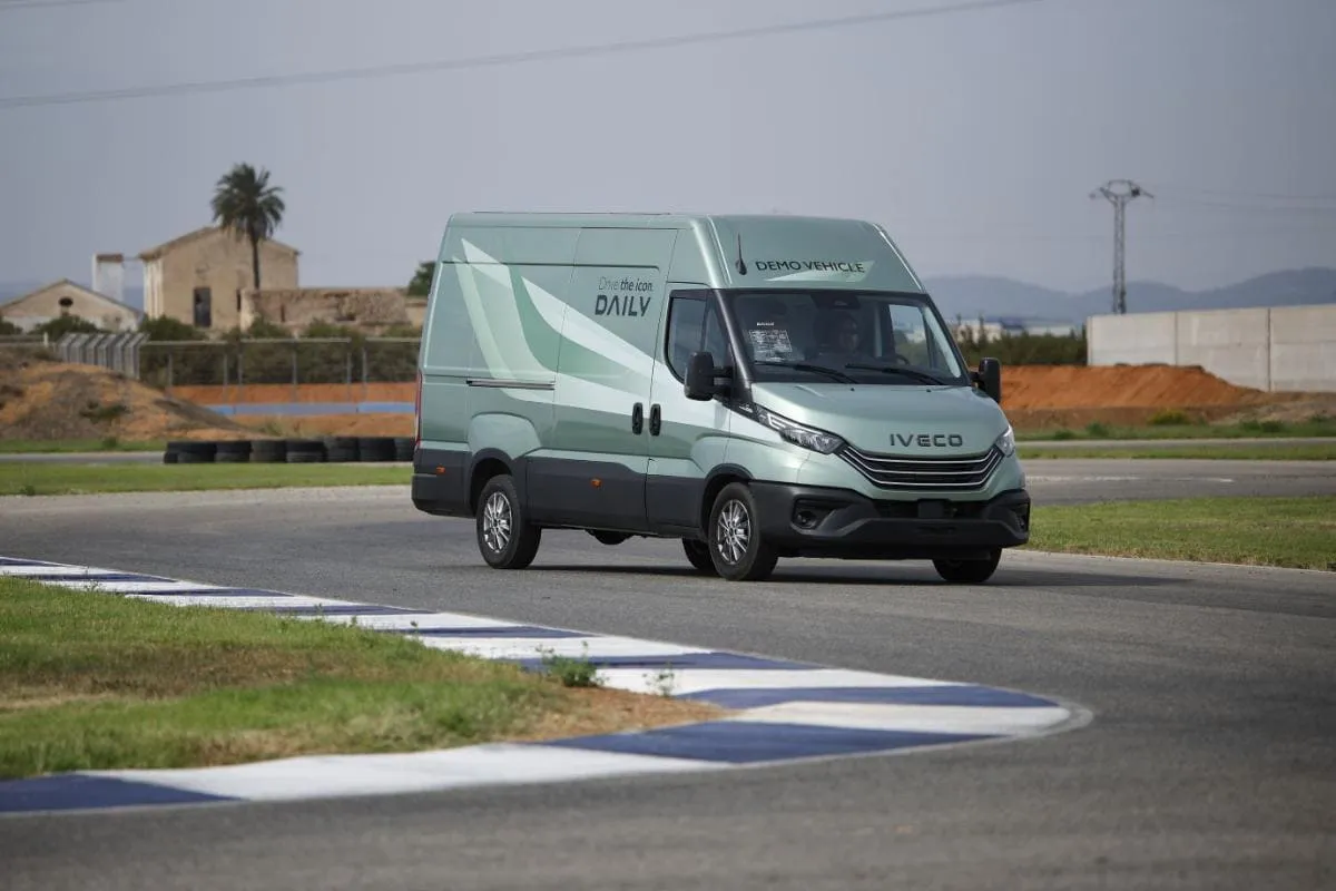 Una furgoneta verde IVECO de la nueva gama circula por una pista flanqueada por edificios y palmeras bajo un cielo nublado en el Circuito Aspar de Valencia.